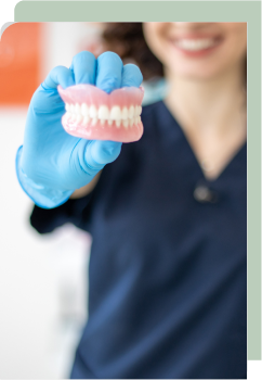 A smiling dental professional holds up a set of dentures with blue-gloved hand, the focus on the dentures with a blurred background.