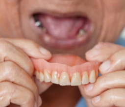 An older person holds a set of upper dentures with one hand, their mouth open and gums visible in the background.