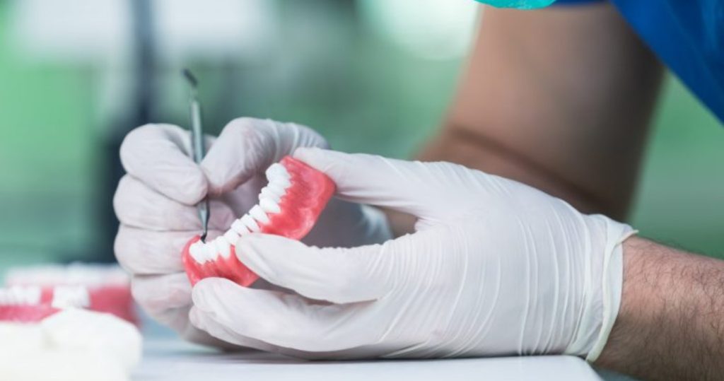 A dental technician wearing gloves and a face mask meticulously works on a set of dentures, using a small tool to make adjustments. The dentures, with a pink base and white teeth, rest on a work surface alongside other dental equipment.