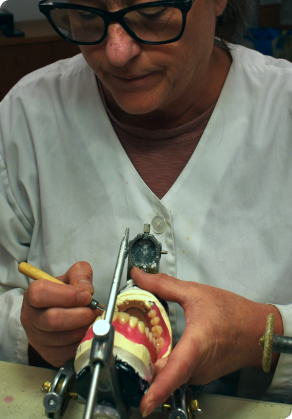 A dental technician in glasses and a lab coat carefully adjusts dentures using precision tools, focusing intently on the dental model.