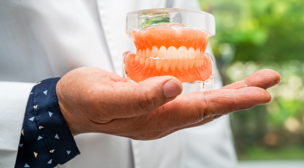 A close-up of a dentist's hand holding a model of dentures, showcasing a full set of artificial teeth, with a blurred green background.
