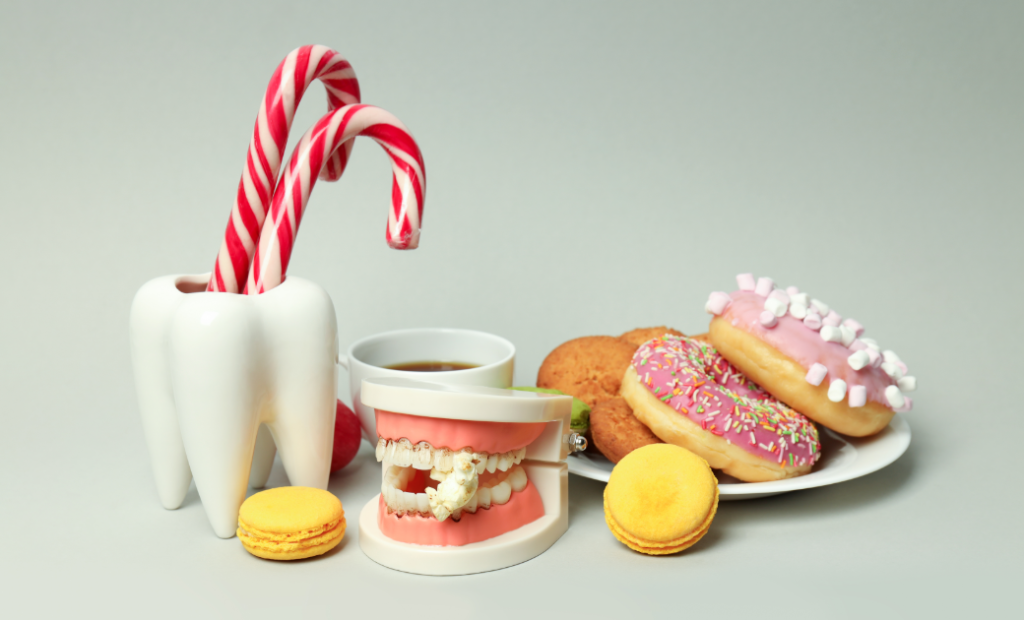 A ceramic tooth holds candy canes next to a dental model biting popcorn. Surrounding them are colorful macarons, cookies, and donuts, illustrating sugary snacks.
