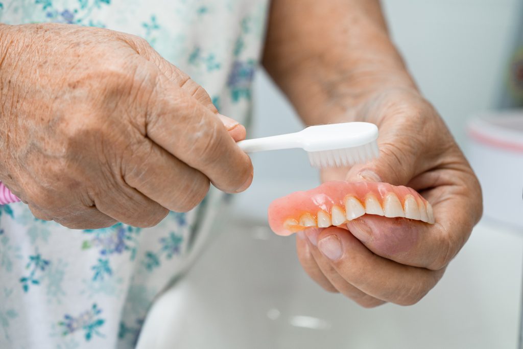 Senior woman cleaning dentures with a soft toothbrush over a sink.
