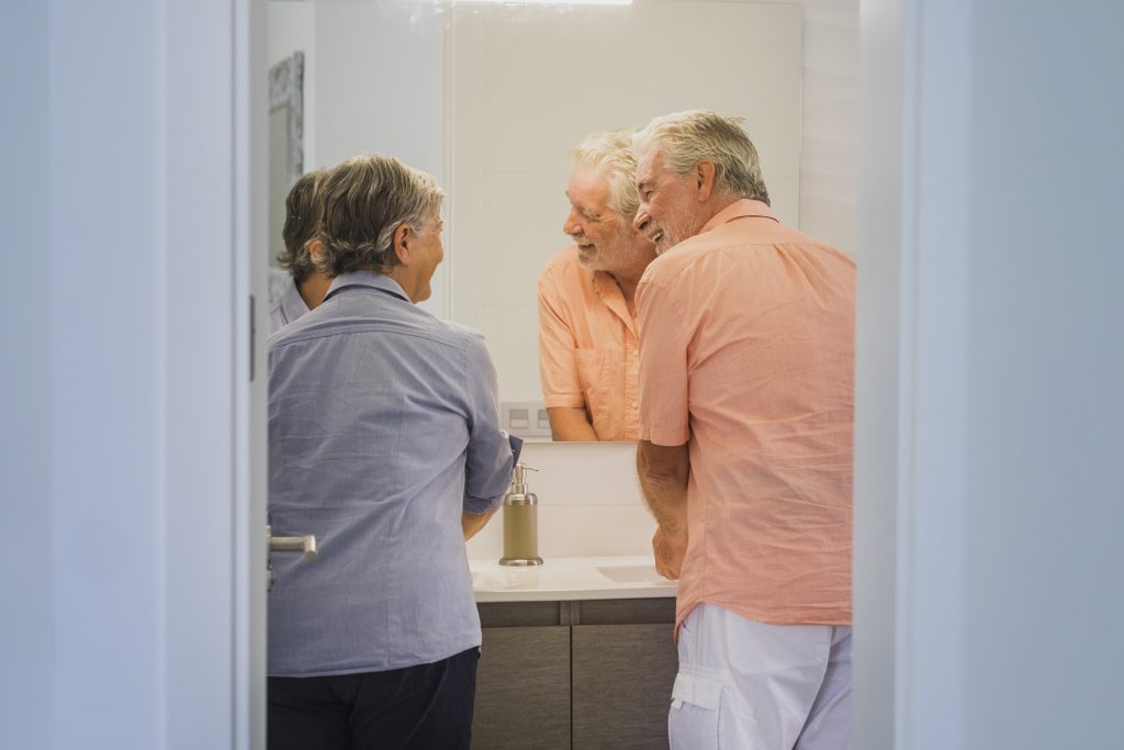 Senior couple smiling in a bright bathroom, gently cleaning dentures together